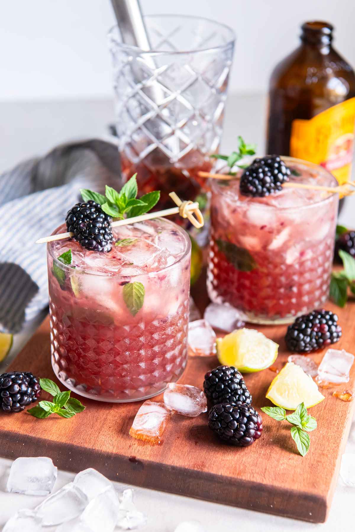 Two blackberry bourbon smash cocktails on a wooden board with fresh blackberries, lime wedges, and mint.