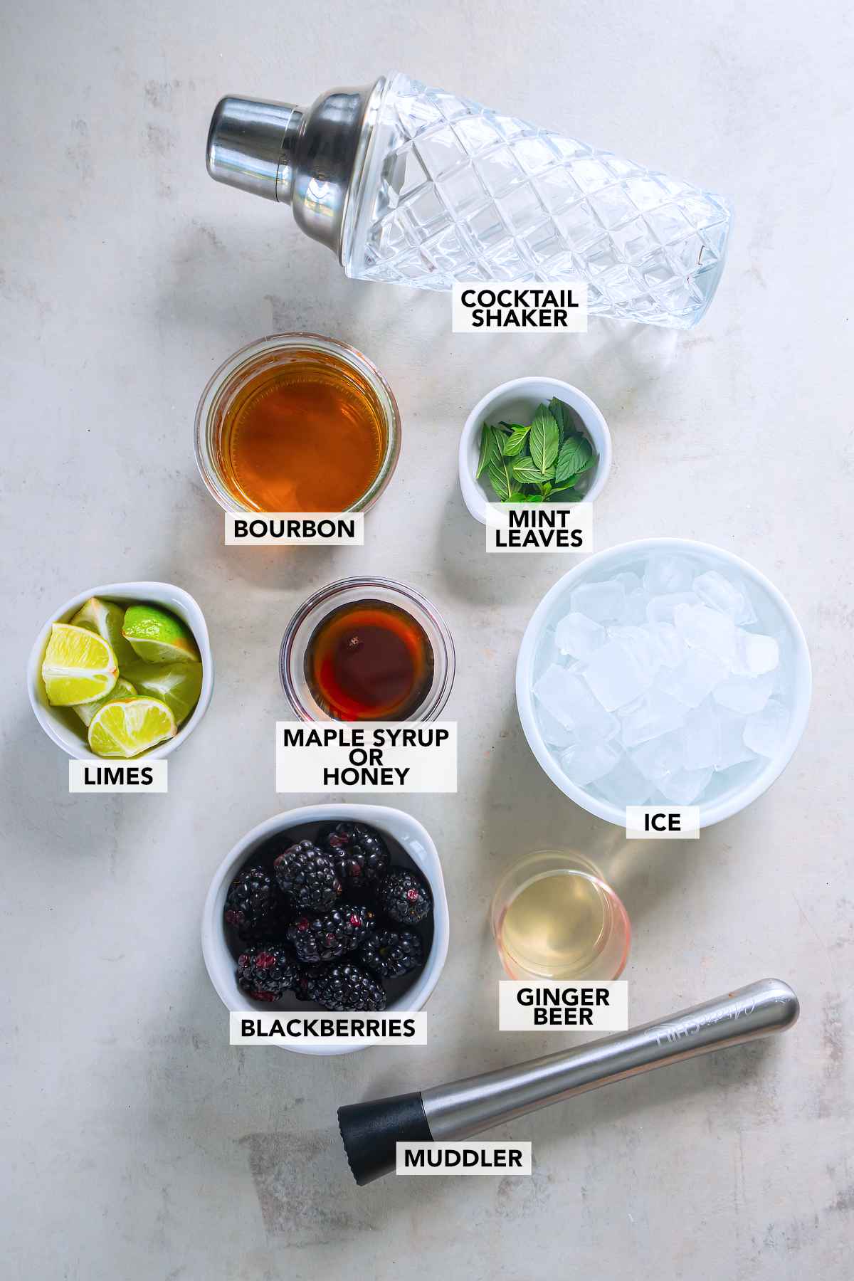 Labeled bourbon, mint, limes, maple syrup or honey, ice, blackberries, and ginger beer beside a cocktail shaker and muddler.