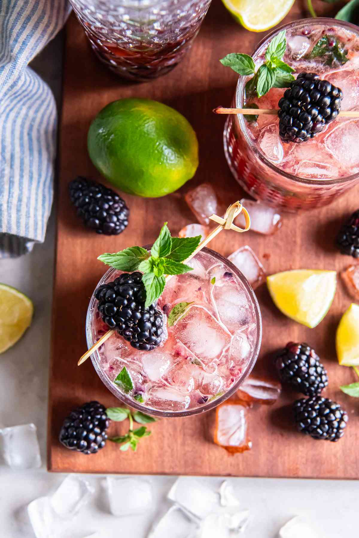 Overhead view of two blackberry bourbon smashes garnished with mint and blackberries, surrounded by lime wedges and ice.