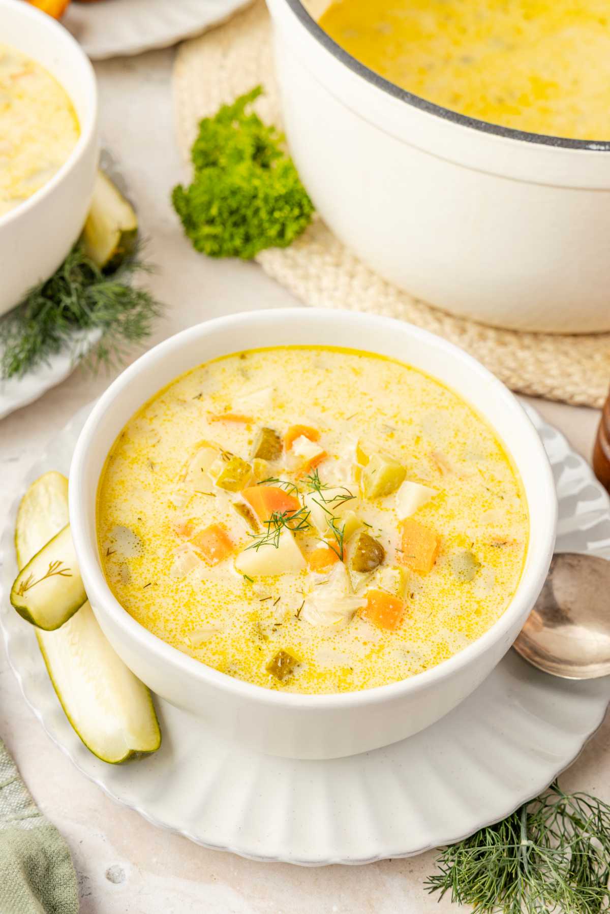 Bowl of creamy dill pickle soup garnished with fresh dill, served beside pickle spears with a pot of soup behind it.