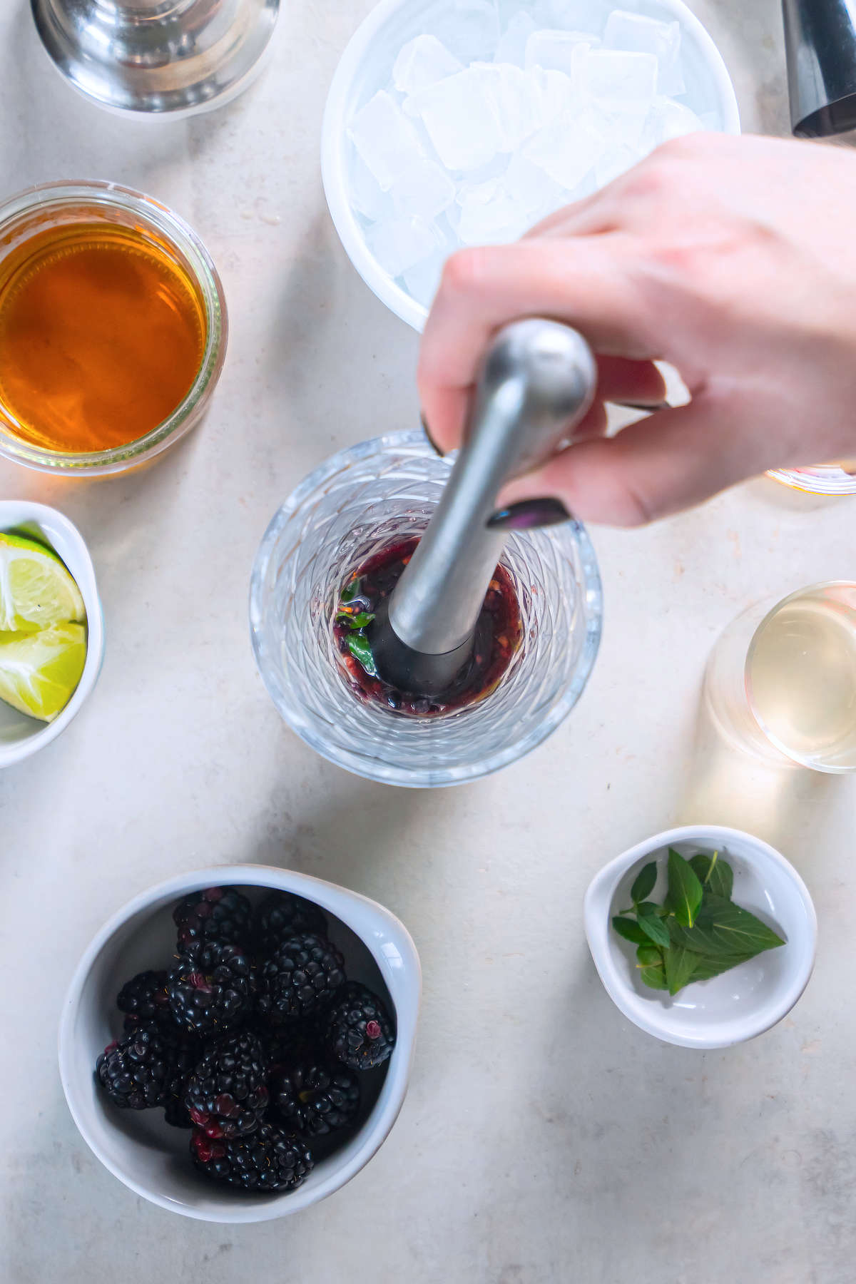 A hand using a muddler to crush blackberries with mint and lime in a glass cocktail shaker.