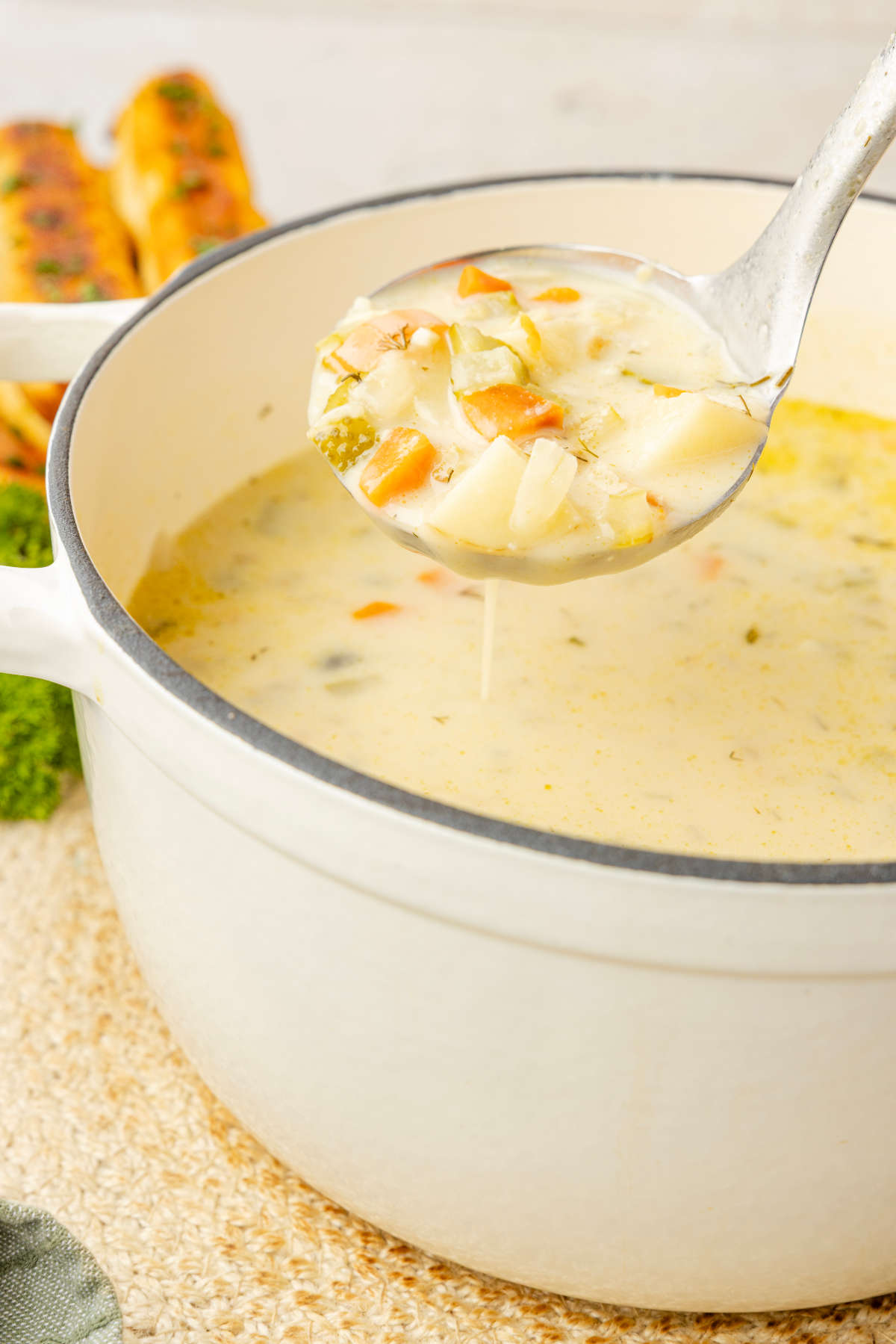 Ladle of creamy dill pickle soup held above a white Dutch oven, with potatoes, carrots and pickles visible.