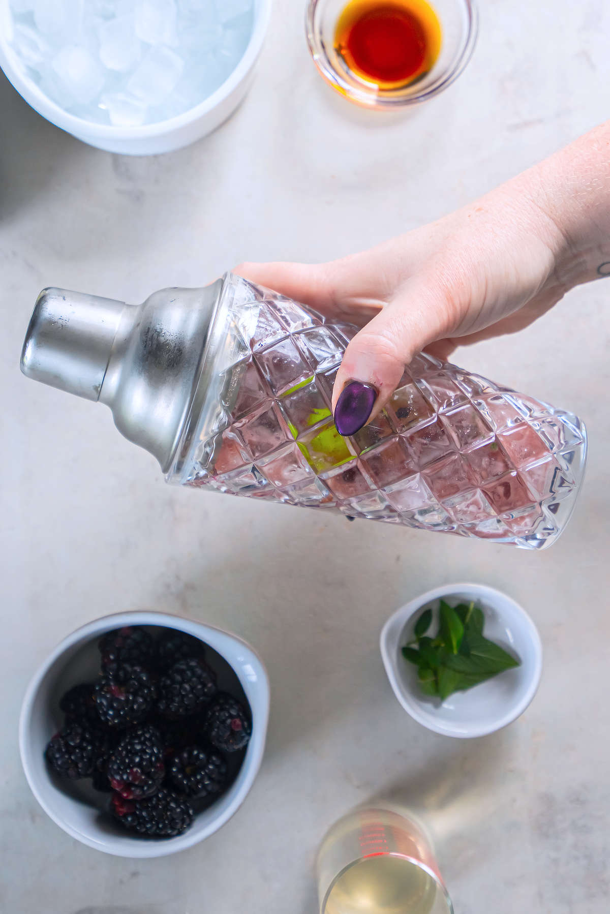 A hand holding a closed glass cocktail shaker with the blackberry bourbon mixture, lime, and ice inside.
