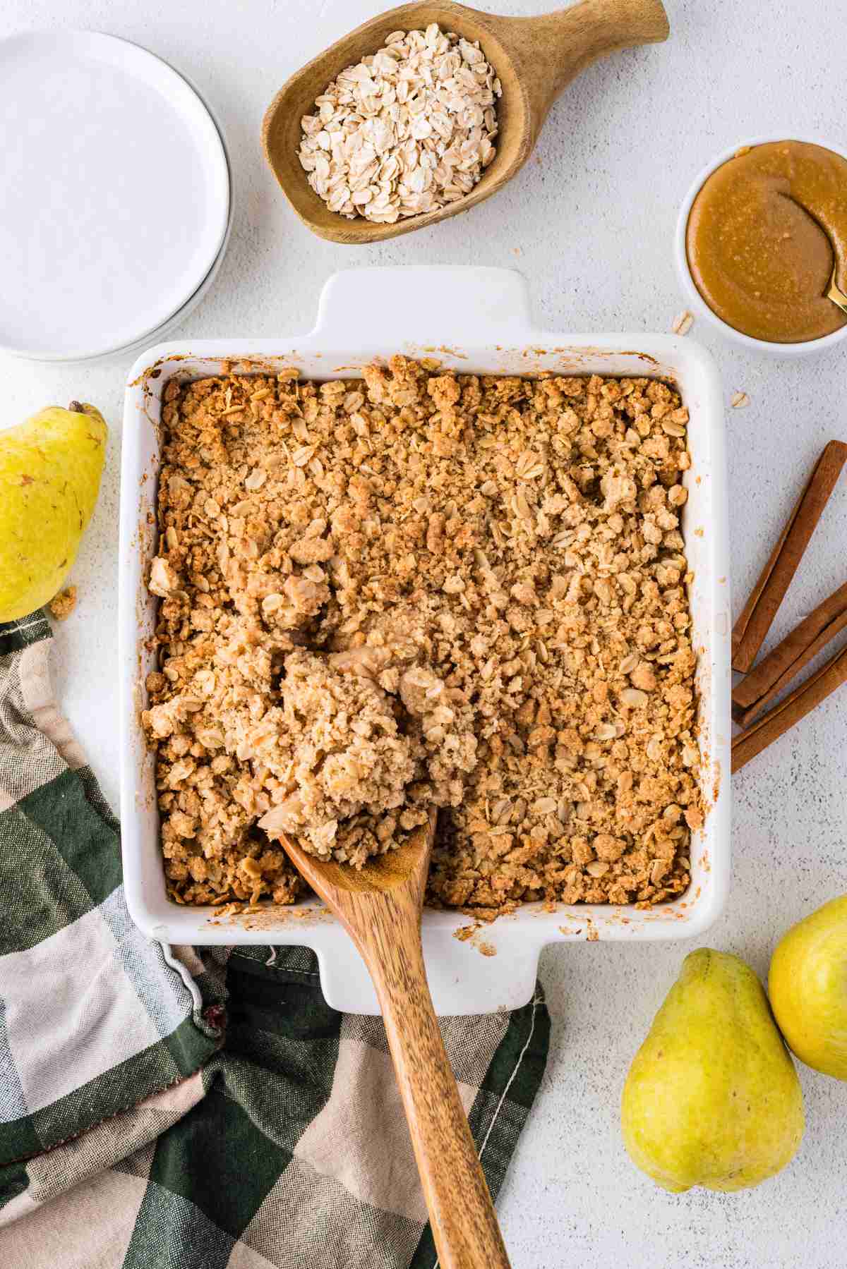 Golden oat topping covering baked pear crisp in a square white dish with a wooden serving spoon.