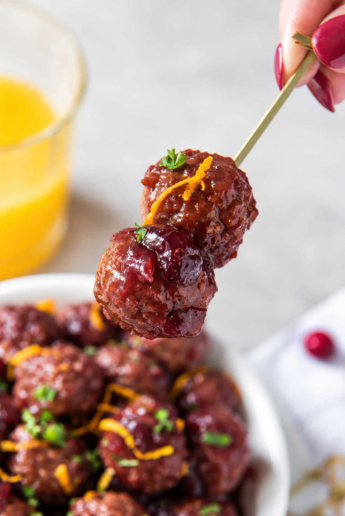 Hand holding a bamboo skewer with two cranberry meatballs coated in glossy sauce and topped with parsley and orange zest.