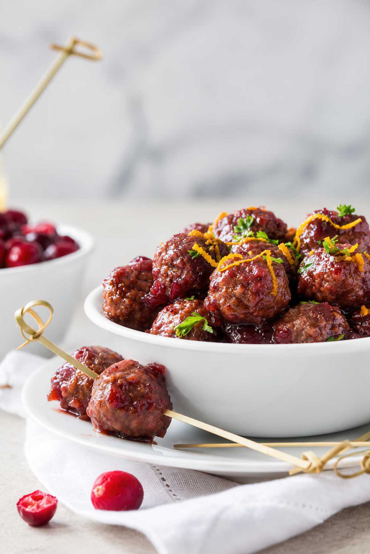Cranberry sauce-coated meatballs in a white serving bowl, with two meatballs on a bamboo skewer in front.