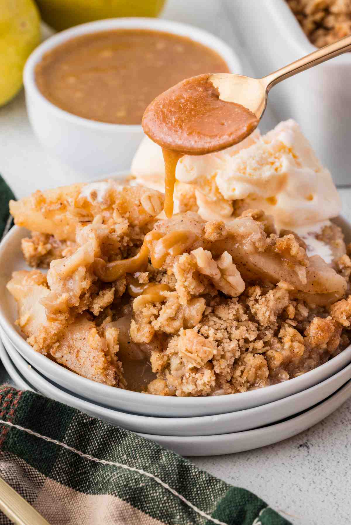 Caramel sauce dripping from a spoon over pear crisp with oat topping and vanilla ice cream.