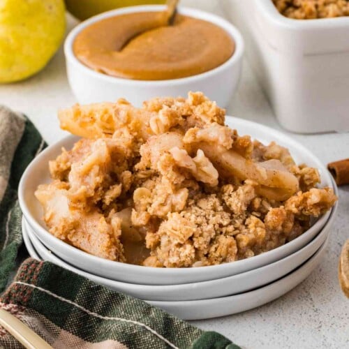 Sliced pears and golden oat crumble piled in a white bowl, with caramel sauce alongside.