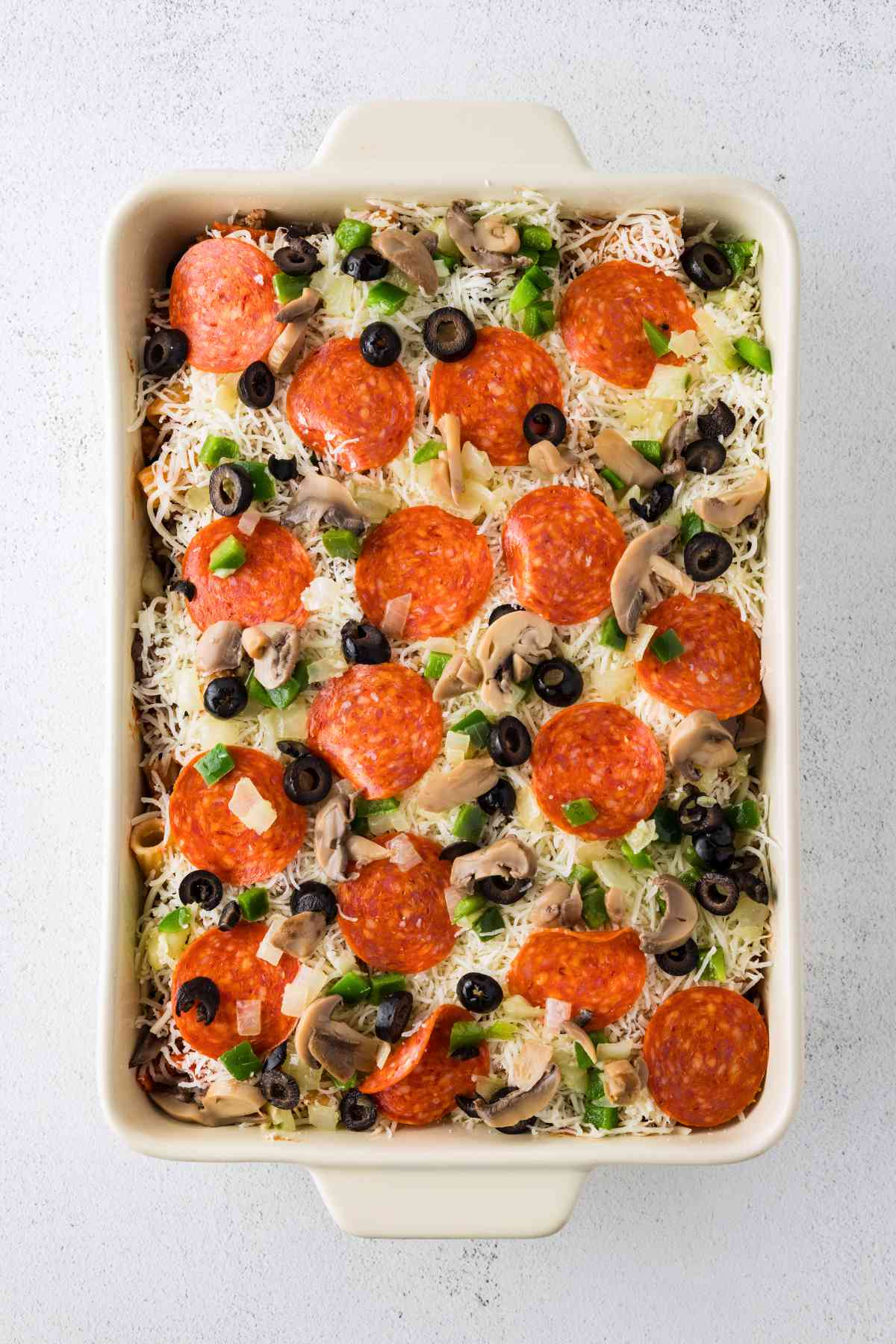 Unbaked pizza casserole topped with shredded cheese, pepperoni, mushrooms, black olives, green peppers, and onions.