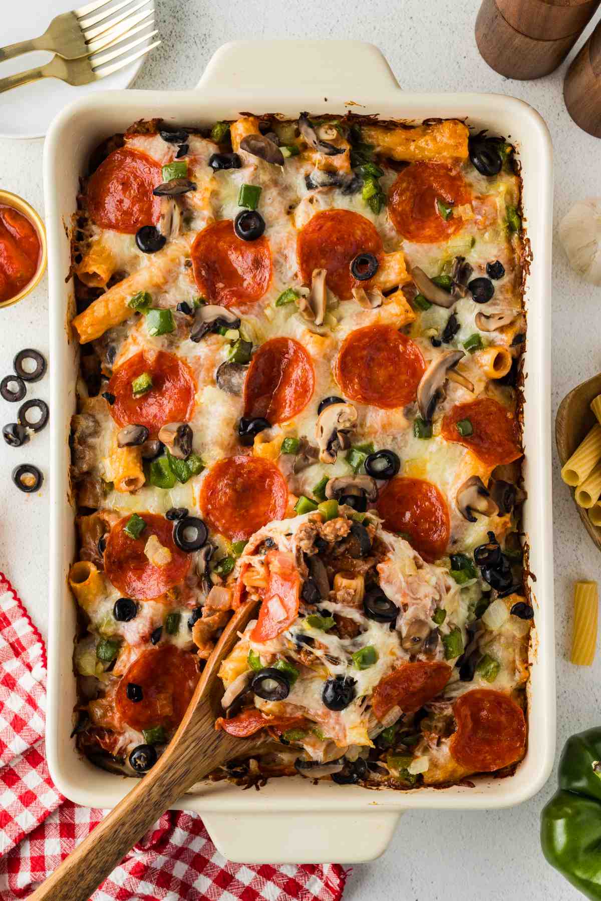 A wooden spoon lifting a serving of pizza casserole from a baking dish topped with pepperoni, melted cheese, and vegetables.
