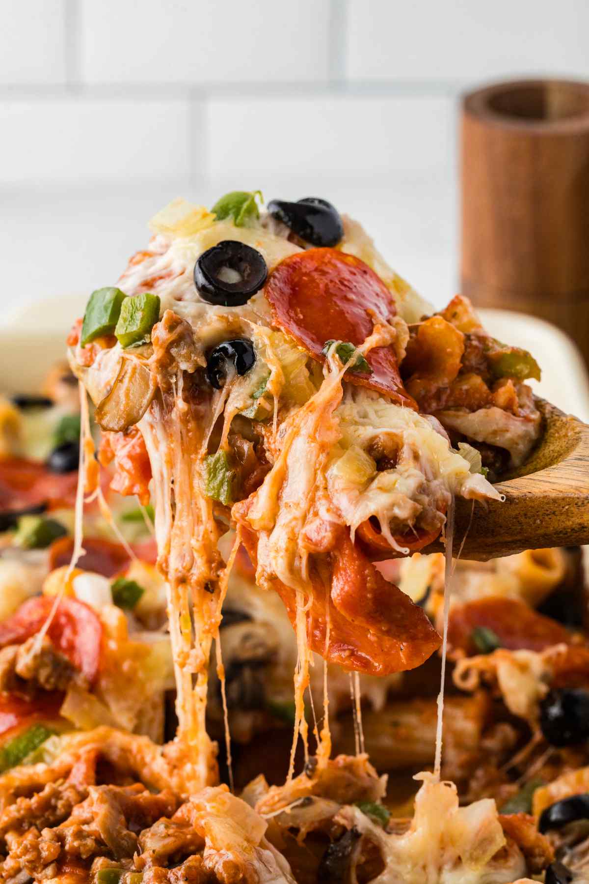 A spoonful of pizza casserole lifted from the dish with strands of melted cheese, pepperoni, black olives, and green peppers.