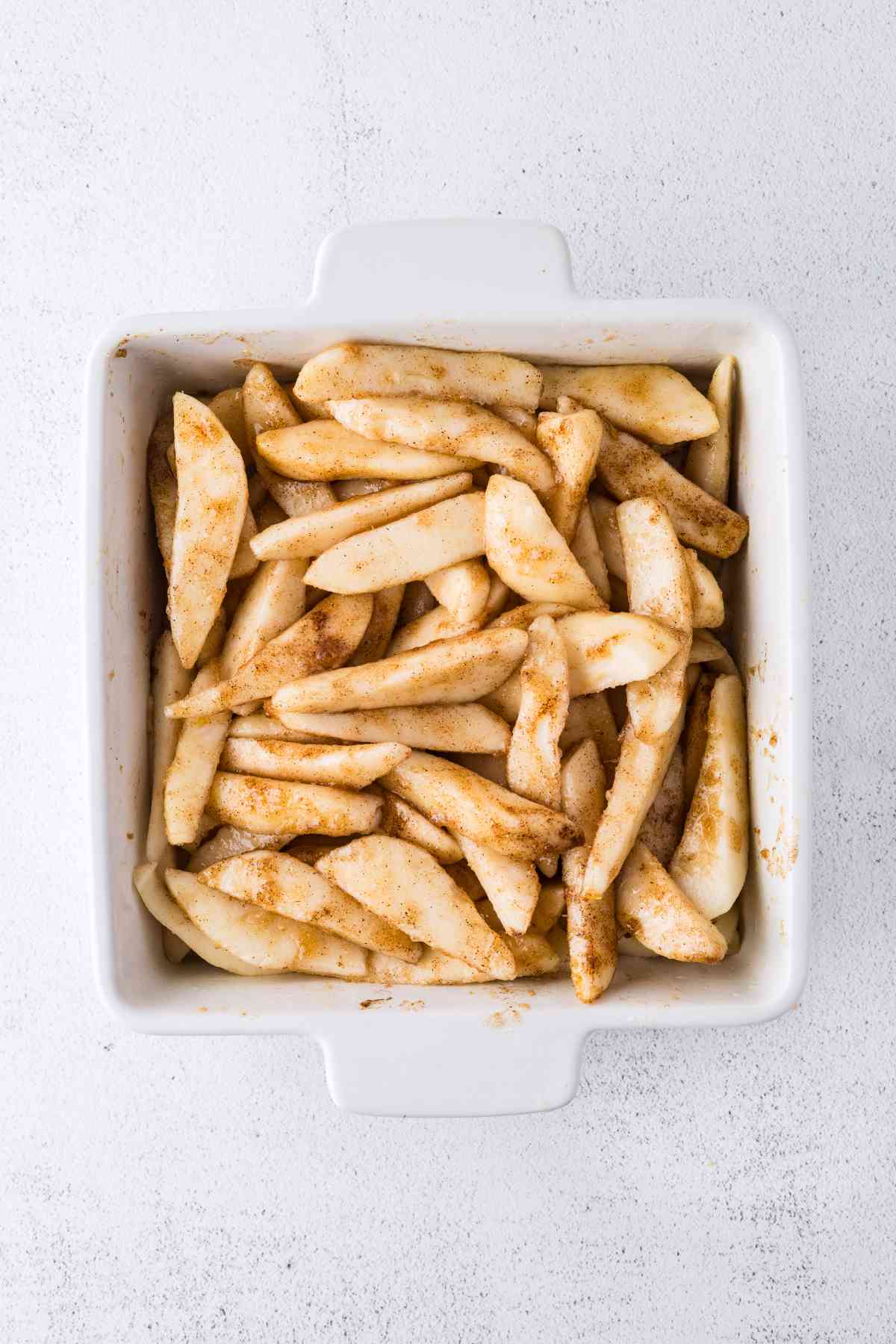 Peeled pear slices coated with brown sugar, cinnamon and nutmeg in a square white baking dish.