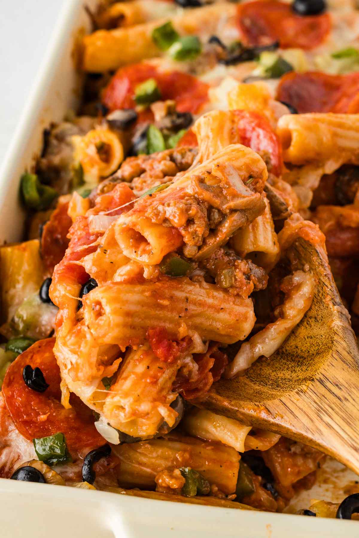 Close-up of rigatoni, sausage, mushrooms, pepperoni, and melted cheese on a wooden serving spoon.