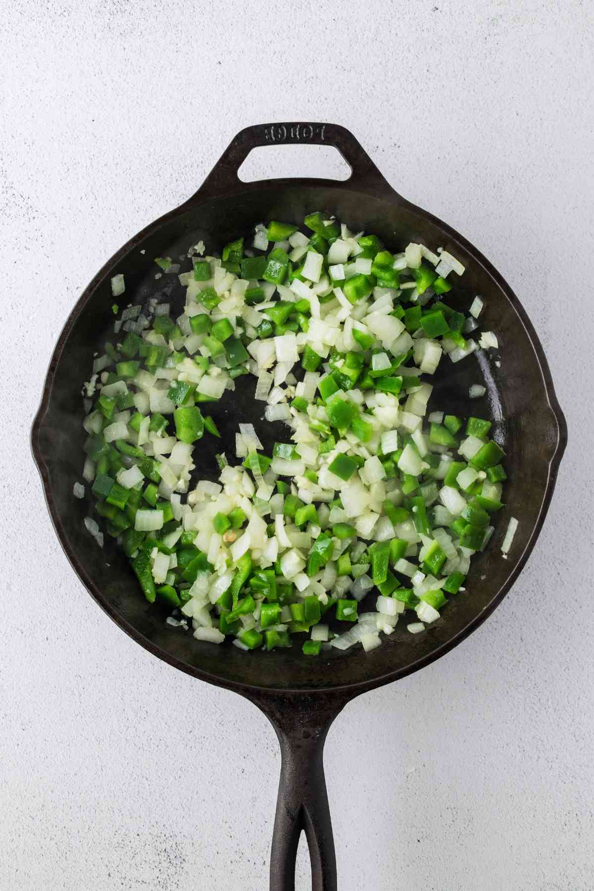 Diced green bell pepper, onion, and minced garlic sautéing in a cast-iron skillet.