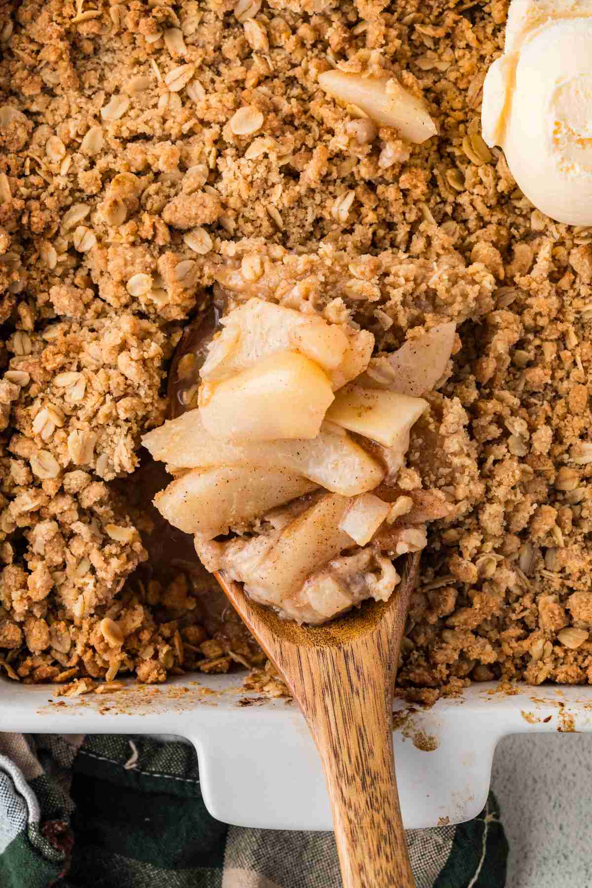 Wooden serving spoon lifting sliced pears and oat topping from a baking dish of pear crisp.