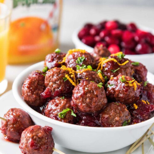 Slow cooker cranberry meatballs garnished with parsley and orange zest in a white bowl, with skewered meatballs on the plate.